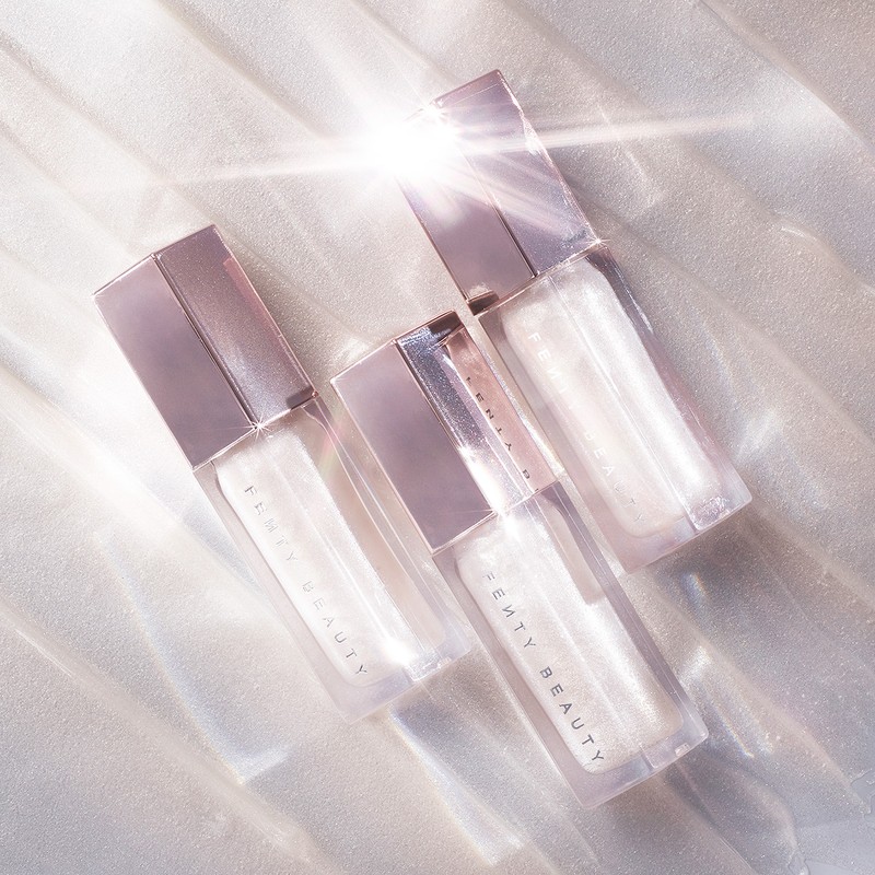 Three Fenty Beauty lip gloss tubes are displayed on a shimmering, white surface with a light flare, highlighting their clear, reflective finish.