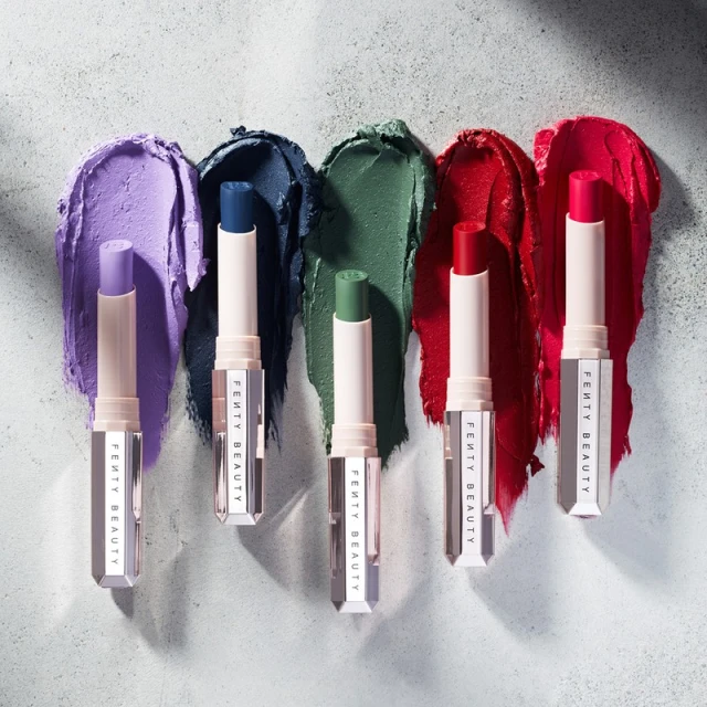 Five Fenty Beauty lipsticks stand against smears of matching color, showcasing shades from purple to red on a textured surface.