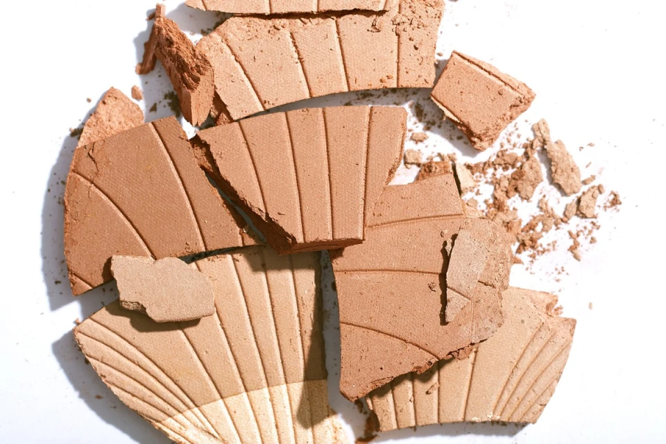 Broken bronzer makeup, close-up overhead shot. Tan and beige shades, shattered powder, on white surface. Beauty and cosmetics imagery.