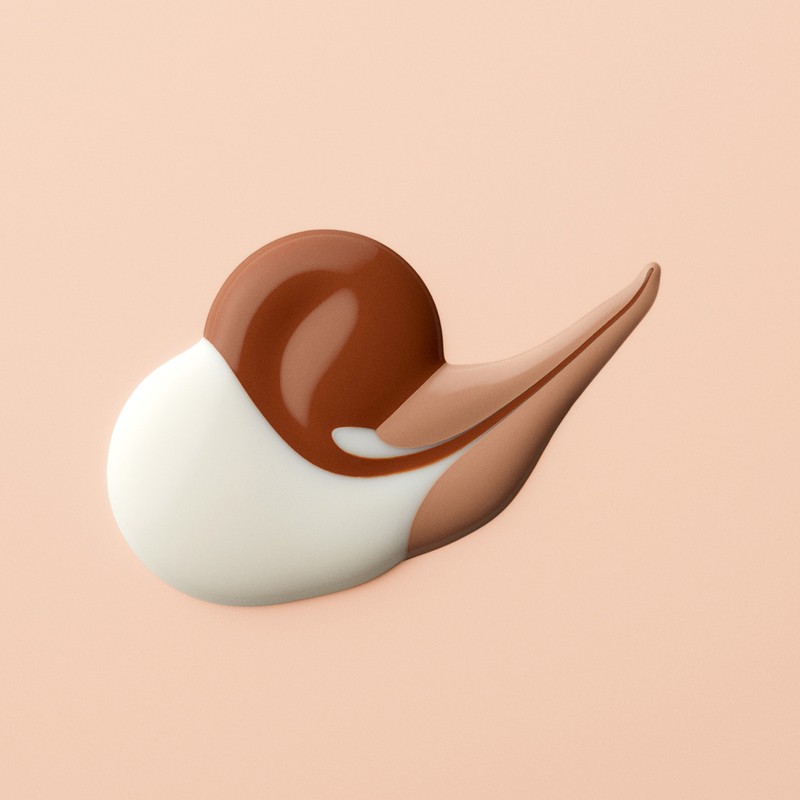 Swirls of brown, tan, and white cosmetic foundation or cream against a light pink background in a beauty product shot.