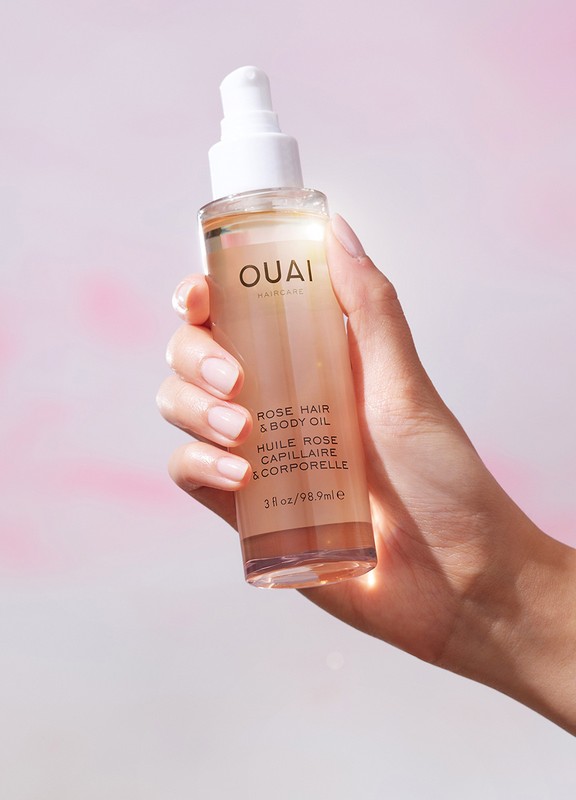 Hand holds bottle of OUAI rose hair and body oil in studio setting with pink background, showcasing beauty and personal care.