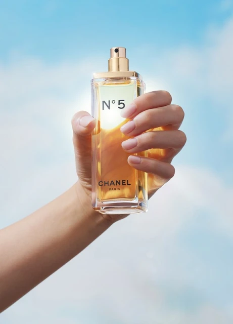 A hand holding a bottle of Chanel N°5 perfume against a blurred sky background, product shot showcasing the gold liquid and glass bottle.