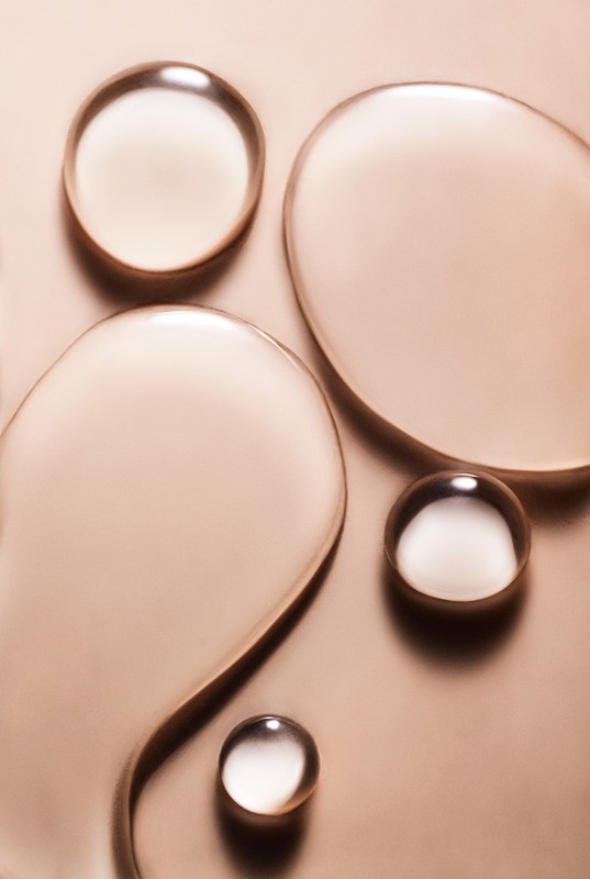 Close-up shot of clear, viscous liquid drops on a metallic bronze background, creating a visually intriguing abstract pattern.