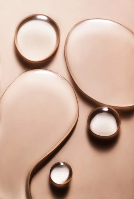 Close-up shot of clear, viscous liquid drops on a metallic bronze background, creating a visually intriguing abstract pattern.