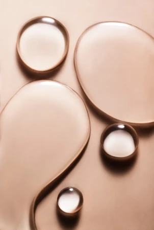 Close-up shot of clear, viscous liquid drops on a metallic bronze background, creating a visually intriguing abstract pattern.