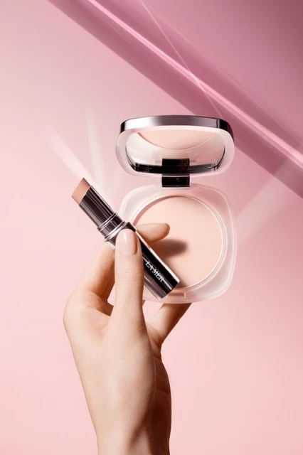 A hand holds a makeup stick next to a compact on a pink background with light streaks, showcasing beauty and cosmetics.