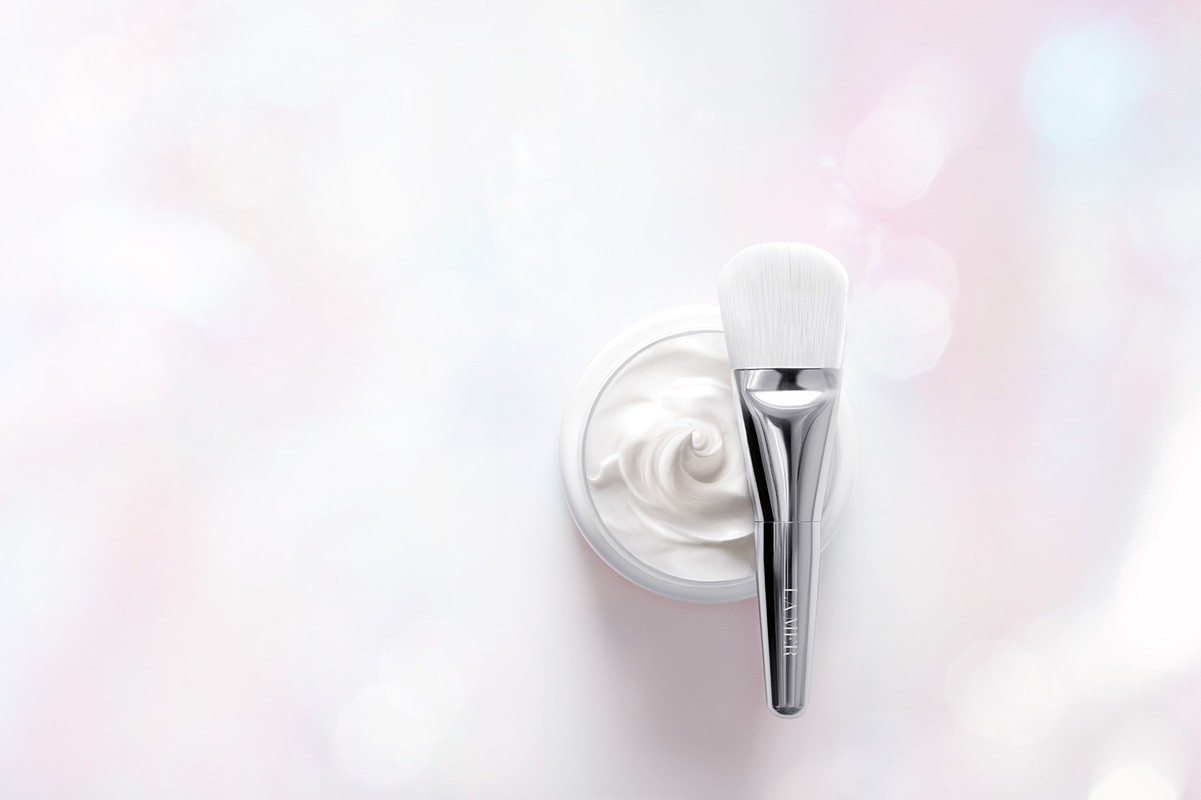 Close-up of a silver makeup brush on a jar of white cream, set against a pastel white and pink bokeh background for beauty shot.