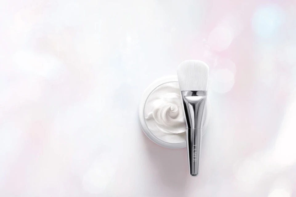 Close-up of a silver makeup brush on a jar of white cream, set against a pastel white and pink bokeh background for beauty shot.