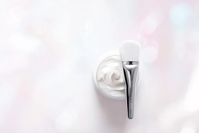 Close-up of a silver makeup brush on a jar of white cream, set against a pastel white and pink bokeh background for beauty shot.