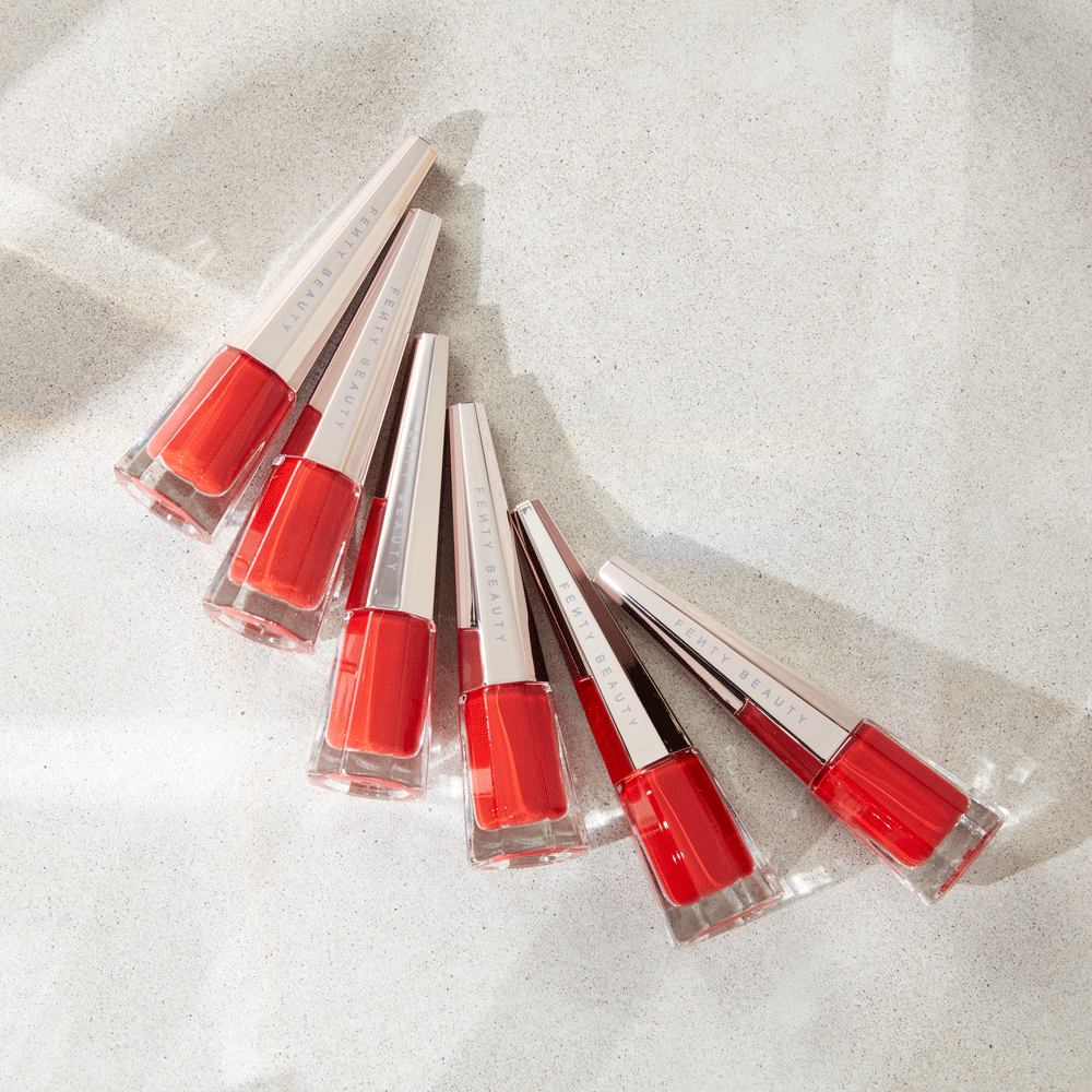 Six Fenty Beauty nail polish bottles with silver caps and vibrant red polish on a light grey textured surface in studio lighting.