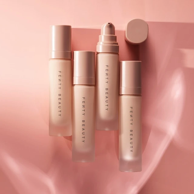 Four bottles of Fenty Beauty foundation in a gradient of shades are arranged on a pale pink surface, casting soft shadows.