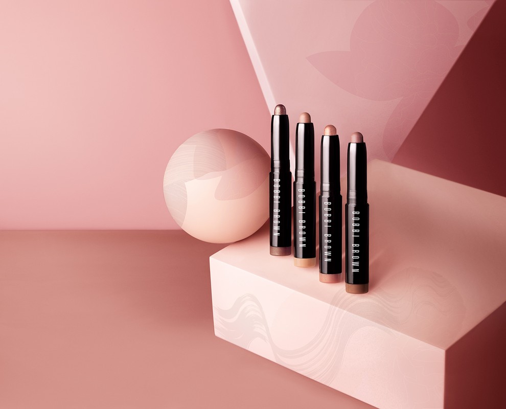 Four Bobbi Brown shadow sticks in neutral tones stand on a geometric platform with a pink backdrop and round accent piece.