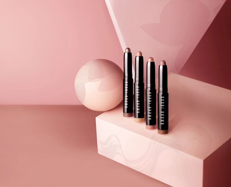 Four Bobbi Brown shadow sticks in neutral tones stand on a geometric platform with a pink backdrop and round accent piece.