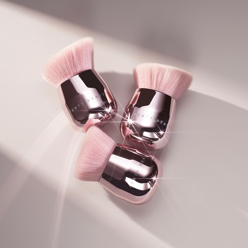Three pink-and-silver Fenty Beauty face and body Kabuki makeup brushes with angled, fluffy bristles on a neutral surface with shadows.