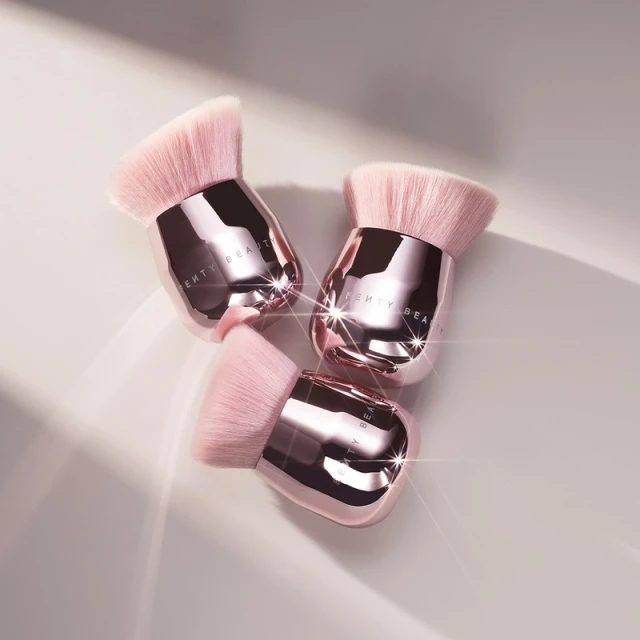Three pink-and-silver Fenty Beauty face and body Kabuki makeup brushes with angled, fluffy bristles on a neutral surface with shadows.