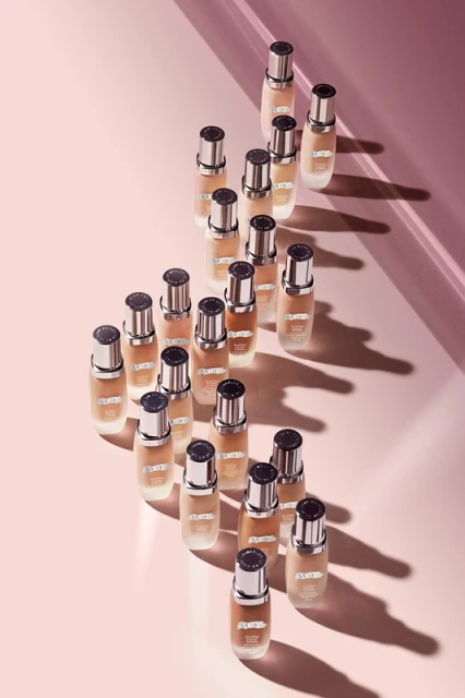 Multiple bottles of La Mer foundation arranged on a pink surface, with shadows adding depth to the beauty product presentation.