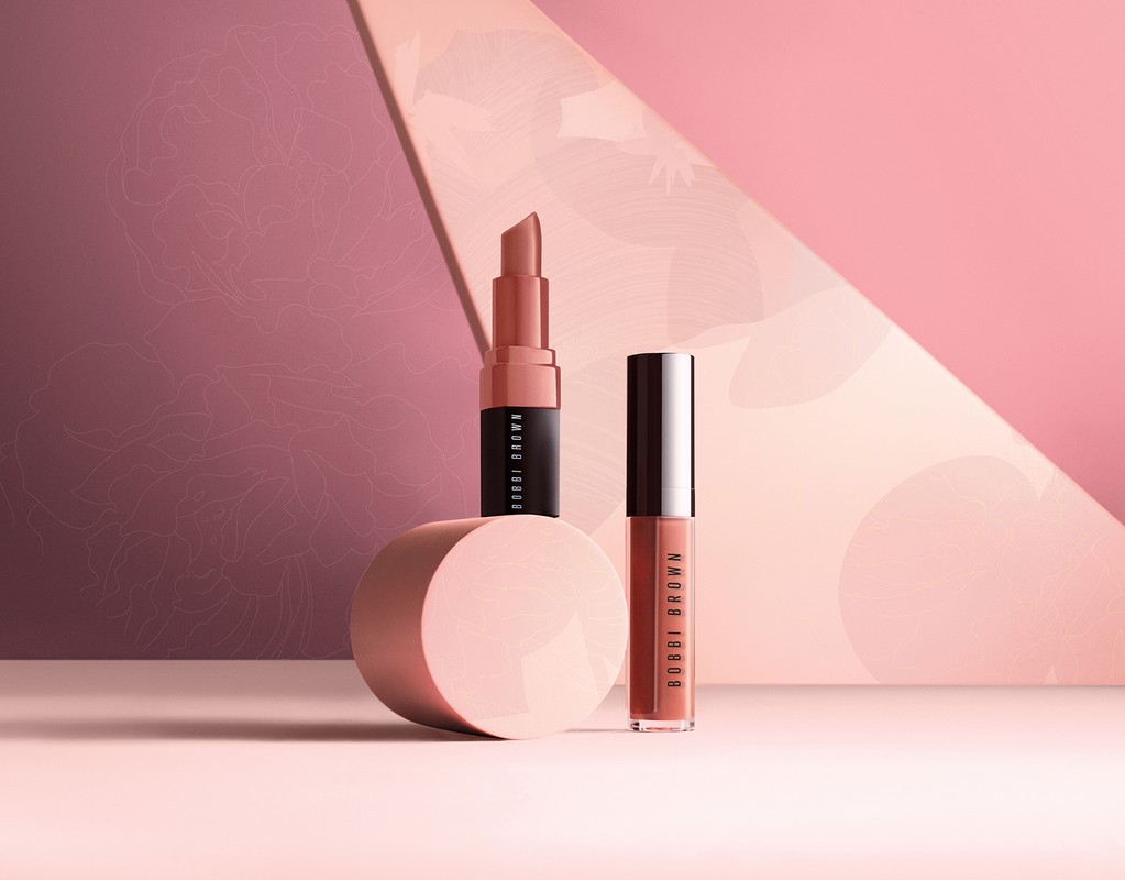 Two cosmetic products, a lipstick and a lip gloss, standing on a pink surface against a geometric two-tone pink background.