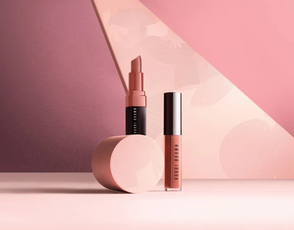 Two cosmetic products, a lipstick and a lip gloss, standing on a pink surface against a geometric two-tone pink background.
