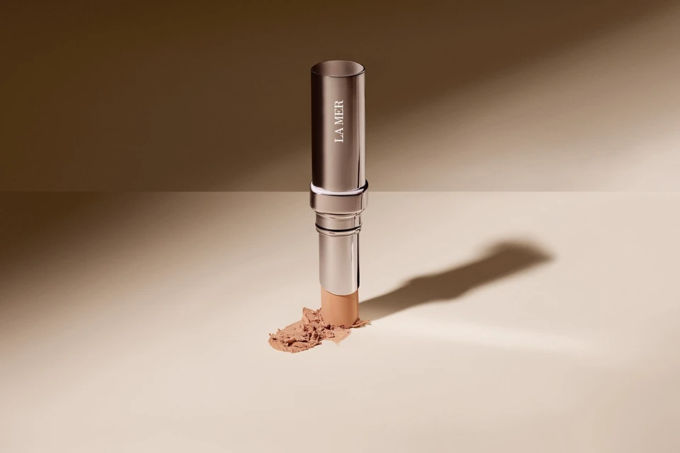Silver La Mer makeup stick with product smudge on beige backdrop, studio lighting, neutral palette, beauty product shot.
