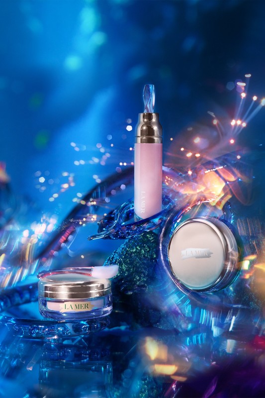 La Mer skincare product assortment on a blue crystalline background with light flares. Cosmetic containers are pink, white, and gold.