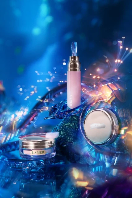 La Mer skincare product assortment on a blue crystalline background with light flares. Cosmetic containers are pink, white, and gold.
