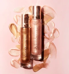 Two tall cylindrical cosmetic bottles with rose gold caps resting on a pale pink background surrounded by shimmering liquid swatches.