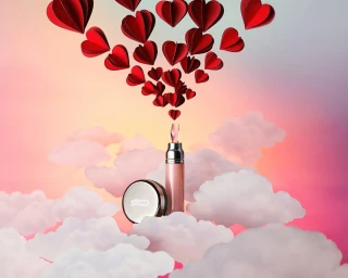 Cosmetics products nestled among pink clouds beneath an arrangement of red paper heart cutouts against a sunset sky.