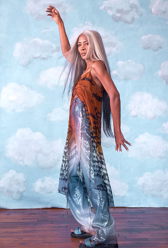 An adult female poses in a studio with cloud backdrop, wearing a colorful sleeveless top, sequined pants and sandals. The model raises her right arm.