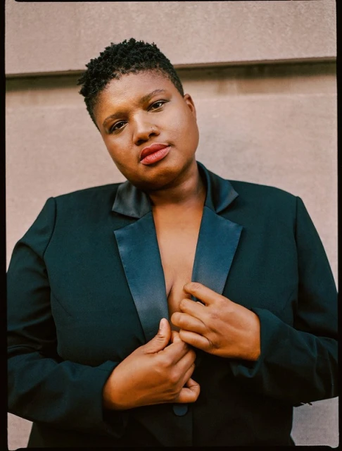 Headshot of an adult female with short black hair wearing a black blazer, looking directly at the camera against a beige wall.