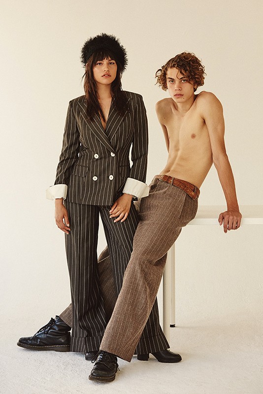 A young couple poses in front of a white background. The woman wears a pinstripe suit and a furry hat, while the man is shirtless.