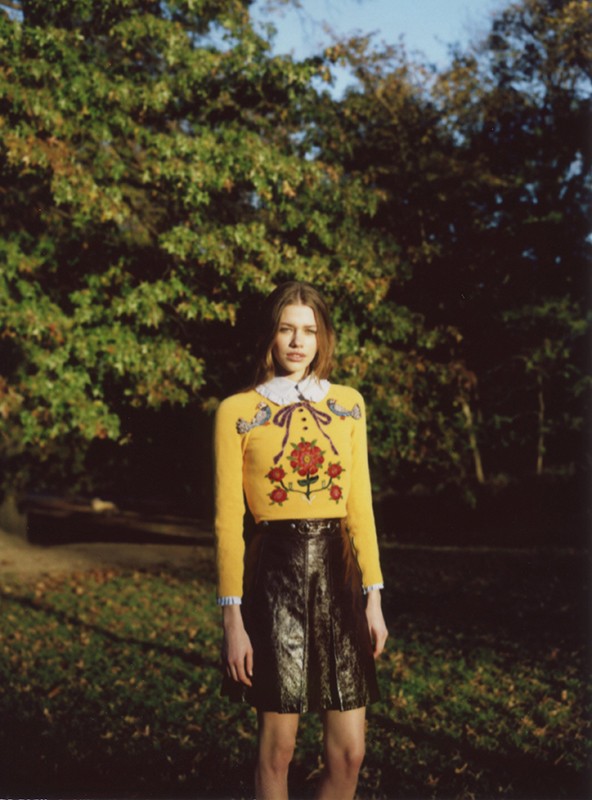 Woman in yellow sweater with rose embroidery and black skirt posing in an outdoor environment surrounded by trees and greenery.