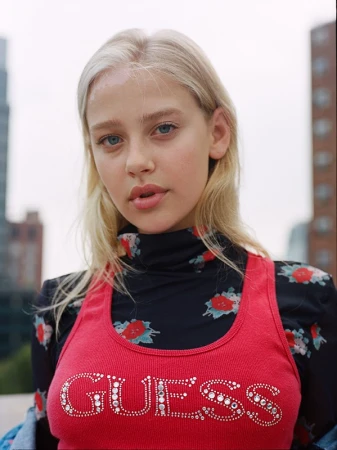 Portrait of a blonde woman wearing a red tank top with rhinestone Guess logo over a black floral turtleneck top.