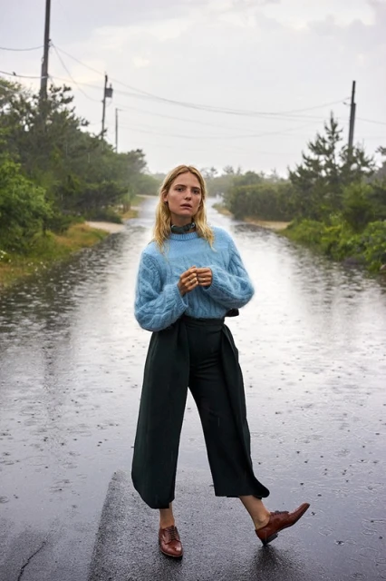 A full shot of a blonde woman in a blue sweater and green pants standing in the rain on a road lined with trees and telephone poles.