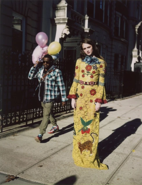 Fashion shot of a model wearing a floral dress with a man holding balloons in the background on an urban street.