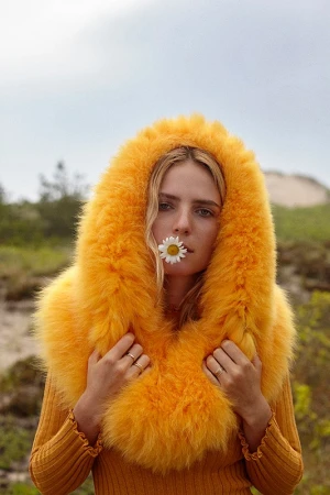 Close-up portrait of a woman wearing a fluffy yellow faux-fur hooded coat holding a white daisy between her lips.
