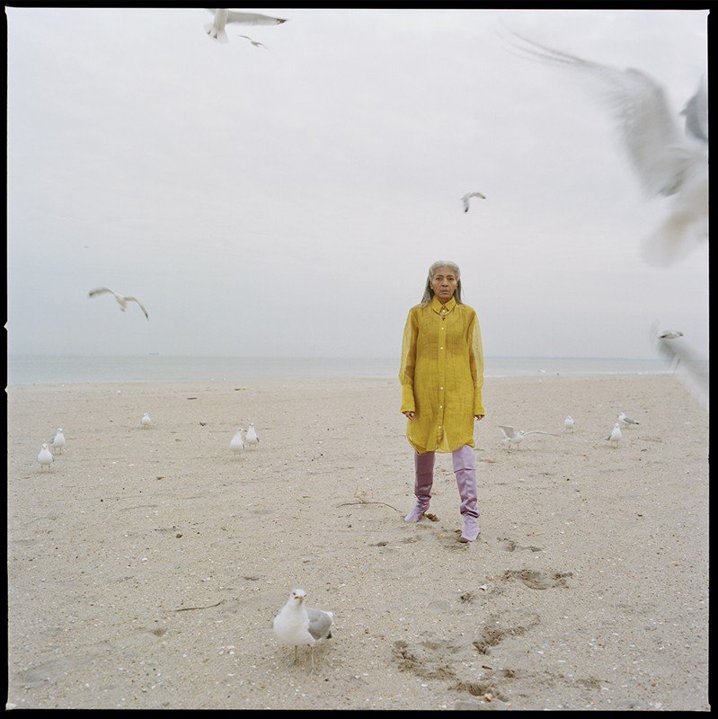 Woman in yellow shirt dress stands on a sandy beach with seagulls, against a pale sky. Wide shot, lifestyle, fashion.