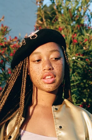 Close-up portrait of a young Black woman with braided hair and freckles, wearing a black beret and satin jacket outdoors.