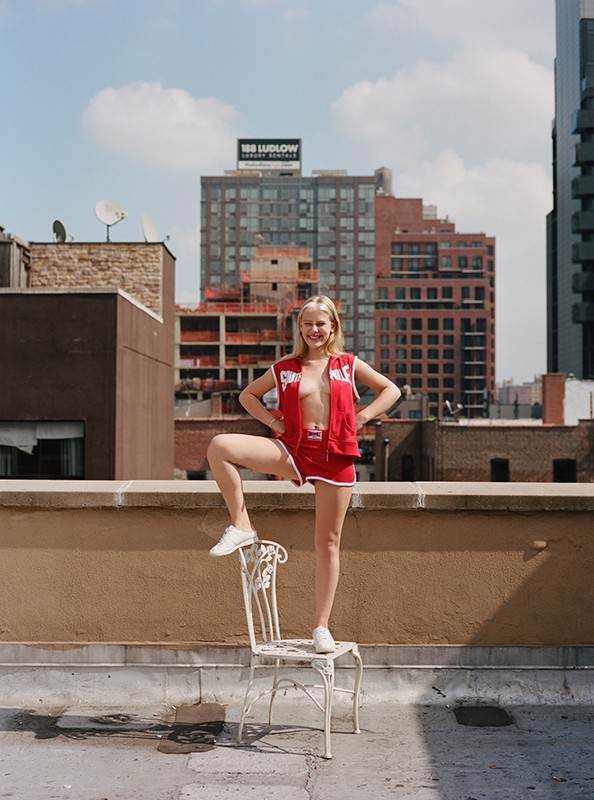 Young woman in a red shorts outfit stands on a white chair on a rooftop with city buildings, clouds, and a blue sky in the background.