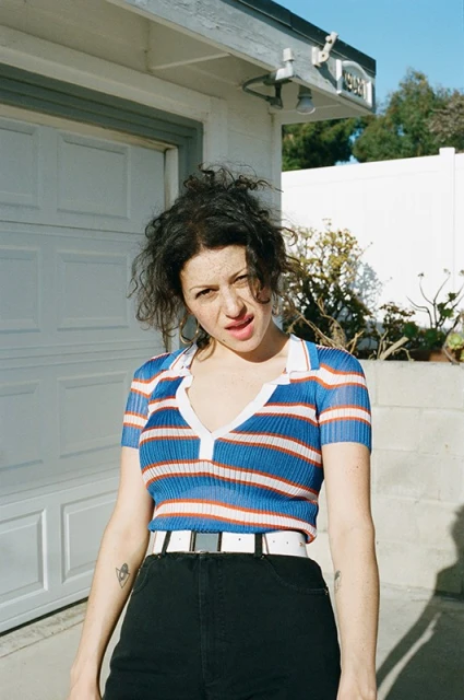 Young woman in a striped collared shirt stands in front of a white garage. The photo is sunlit, highlighting the outdoor setting.