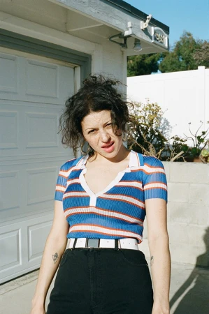 Young woman in a striped collared shirt stands in front of a white garage. The photo is sunlit, highlighting the outdoor setting.