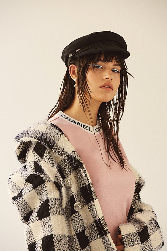 Woman in black hat and checkered coat stands against a white background, wearing a pink shirt, her wet hair framing her face.