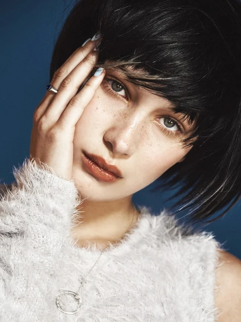 Portrait of a young woman with dark hair and freckles, wearing a fuzzy sweater and silver necklace, against a solid blue background.