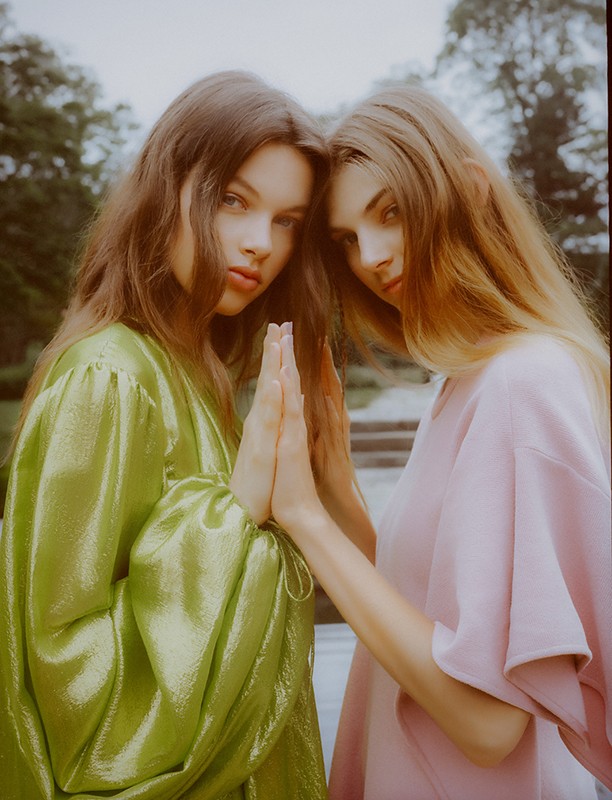 Two female models posing with palms touching, wearing green and pink dresses outdoors styled by Doria Santlofer.
