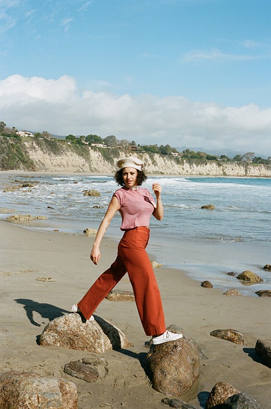 A woman wearing a beret, top, and trousers poses on rocks at a beach. Waves are breaking on the shore on a sunny day.