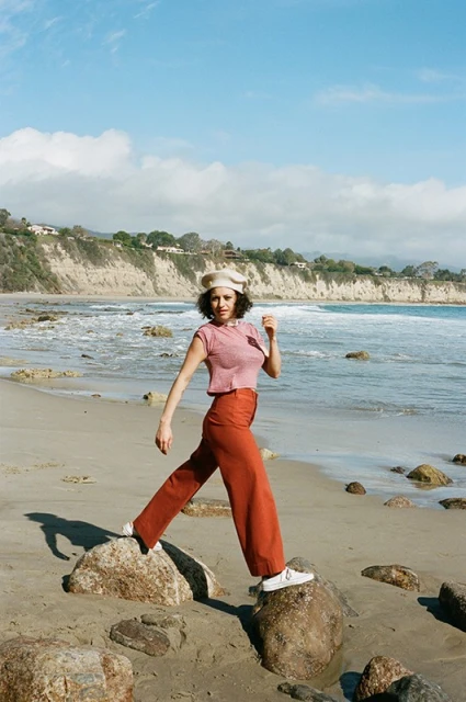 A woman wearing a beret, top, and trousers poses on rocks at a beach. Waves are breaking on the shore on a sunny day.