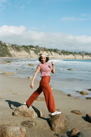 A woman wearing a beret, top, and trousers poses on rocks at a beach. Waves are breaking on the shore on a sunny day.