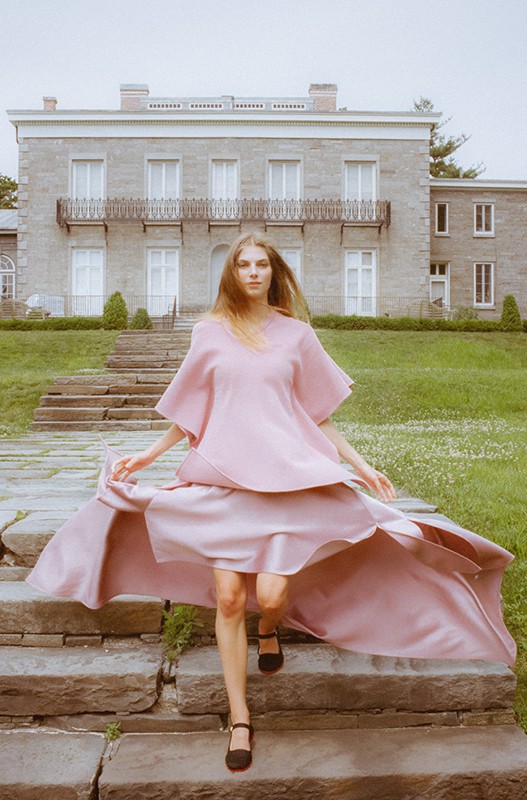 Woman in flowing pink dress descends stone steps in front of large stone building and green lawn on overcast day.