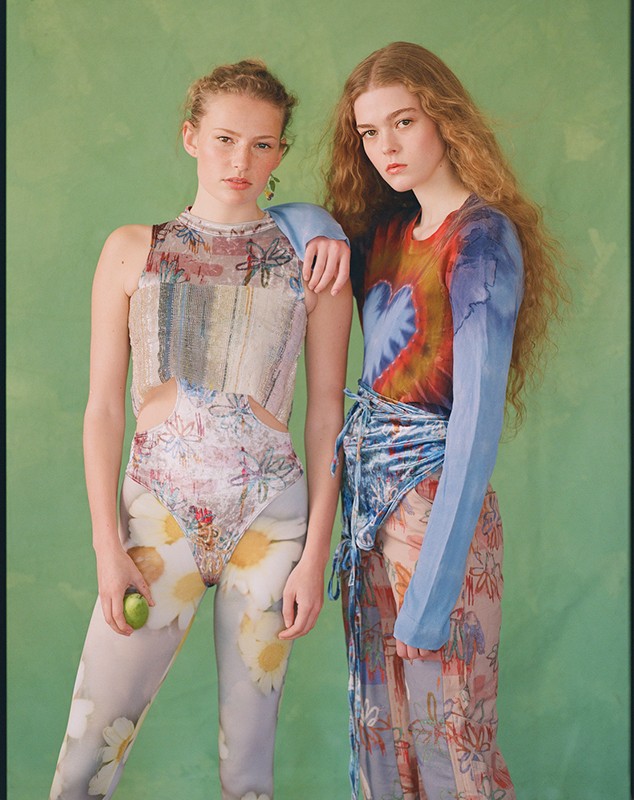 Two fashion models pose in an indoor studio setting with a solid green background. One model holds a green fruit. They wear colorful outfits.