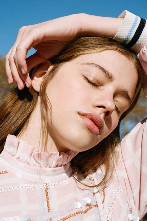 Close-up portrait of a young woman with closed eyes wearing a pink patterned top with a high ruffled collar under a clear blue sky