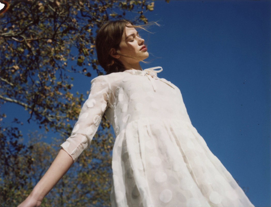 Young woman in a white dress with polka dots stands with arms outstretched, gazing upward into the bright blue sky with dappled sunlight.